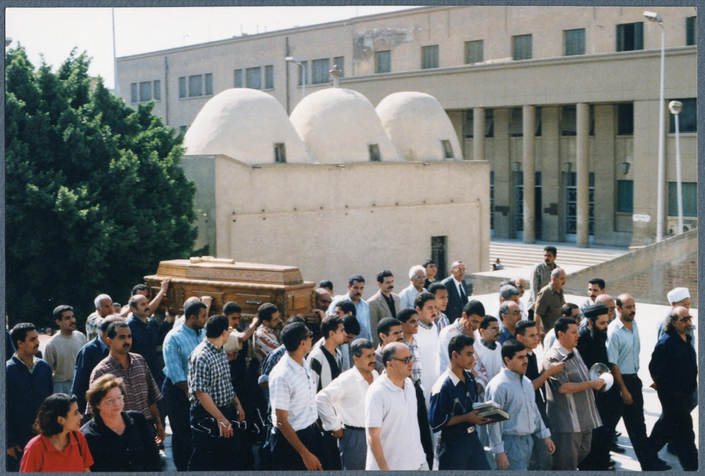 A crowd of men follows a coffin carried through a churchyard; one of them holds a cymbal