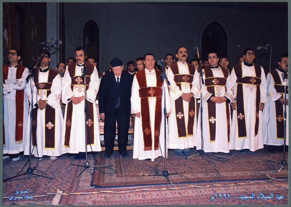 A row of deacons in white vestments with red stoles stands at microphones, with an elderly man in a dark suit among them