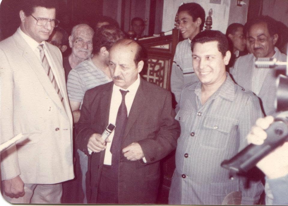 A man in a dark suit and tie holds a small microphone in a church, with men in suits and shirts around him and a camera at the right edge