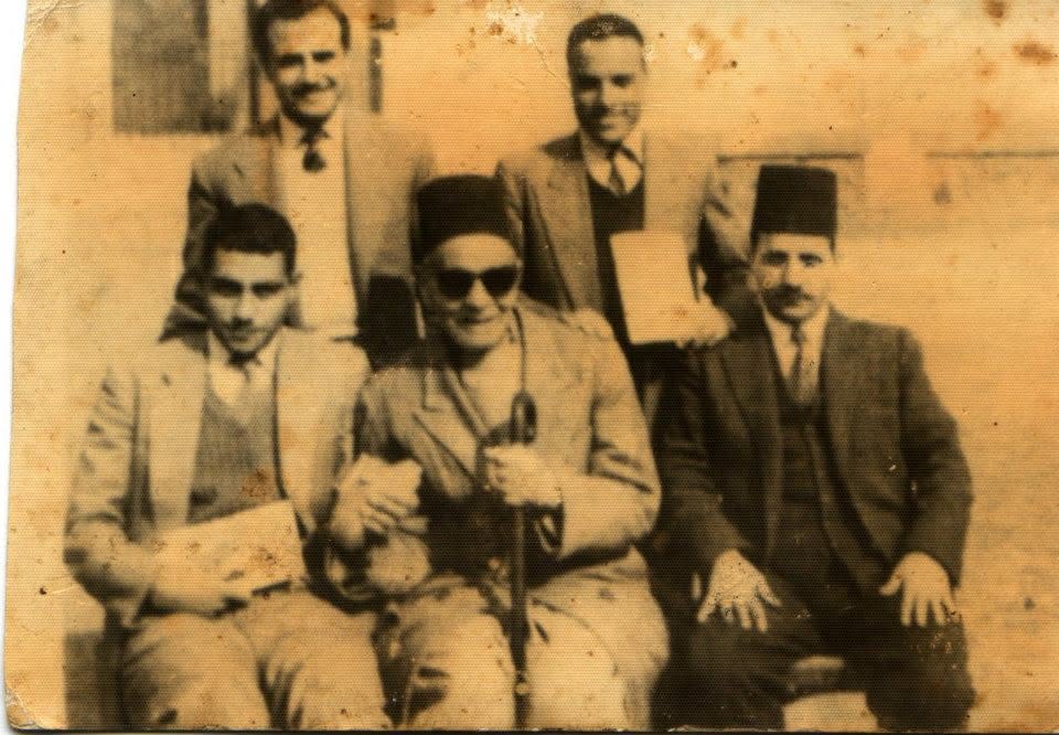 A faded sepia photograph of three men seated in front and two standing behind them, the man in the centre in dark glasses and a tarbush with a cane