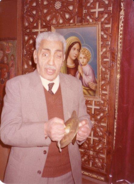 An elderly man in a grey jacket and brown waistcoat holds a pair of cymbals before an icon of the Virgin and Child