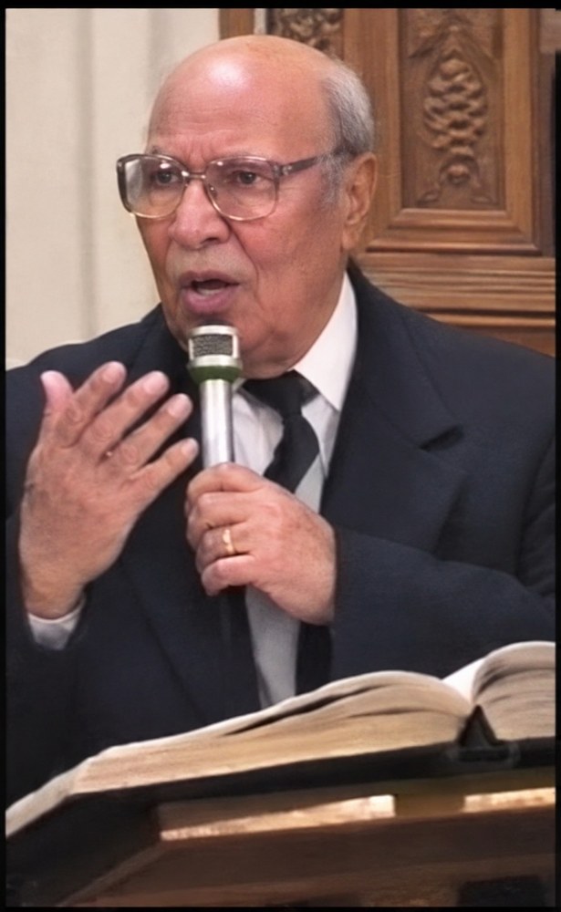 An elderly man with white hair and glasses, in a dark suit, chants into a microphone with an open book before him