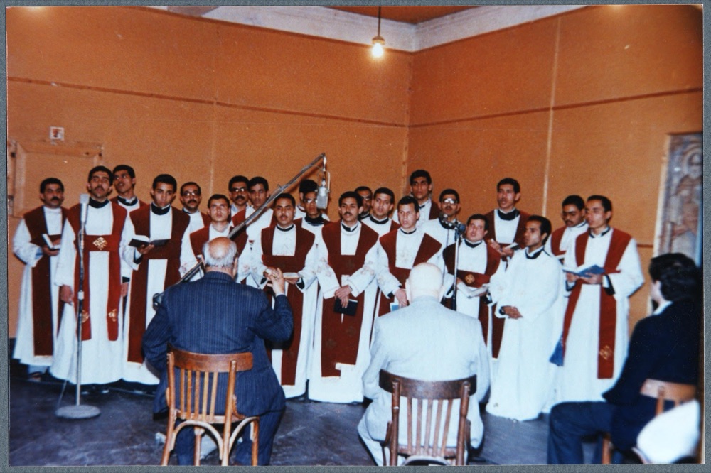 A choir of deacons in white and red vestments sings at a microphone while two men sit before them with their backs to the camera