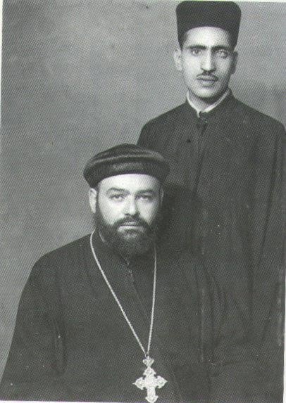 A young man in a black cassock standing behind a bearded priest in a black cap with a pectoral cross