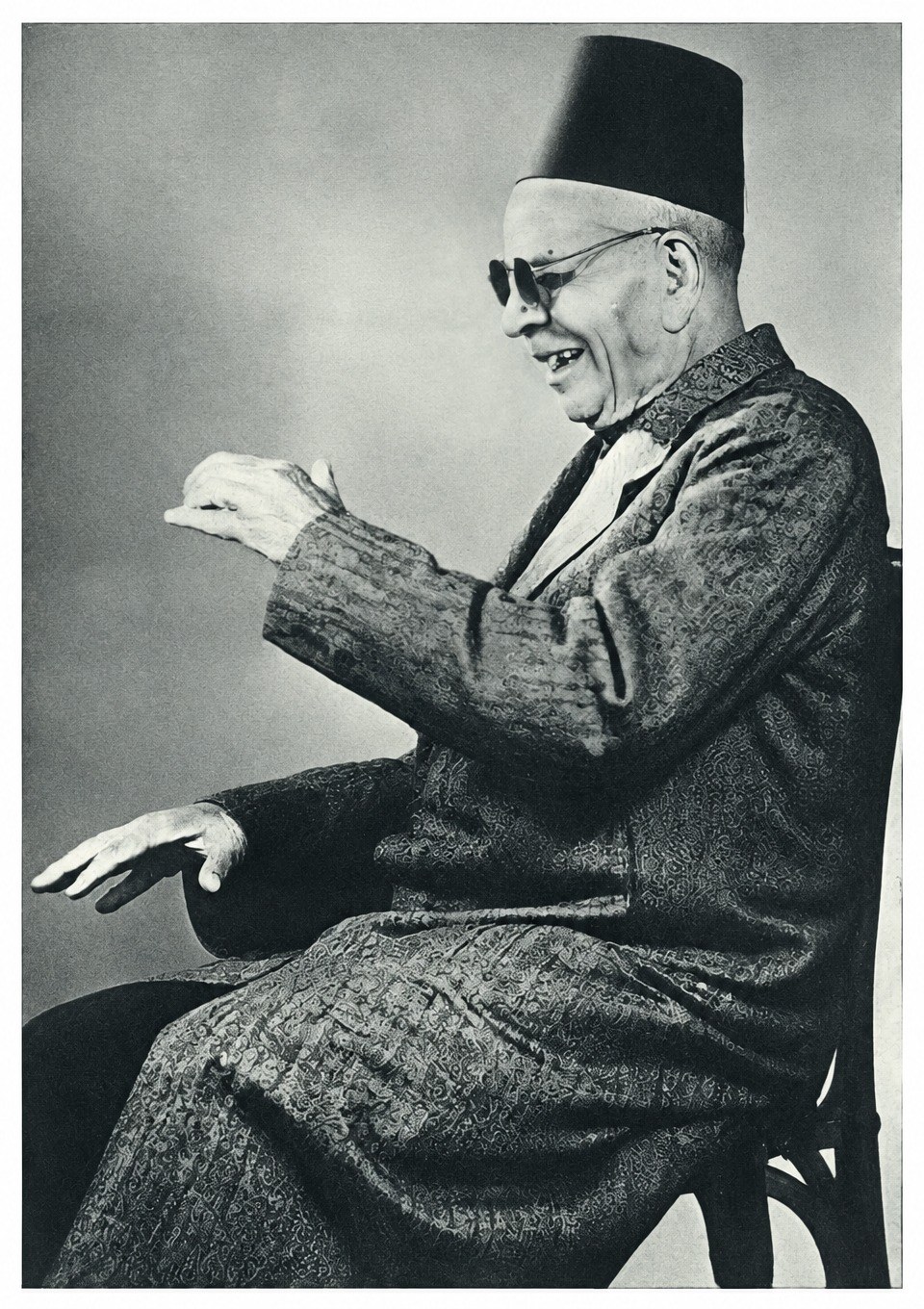 A smiling old man in dark glasses, a tarbush and a patterned robe, seated in profile with one hand raised