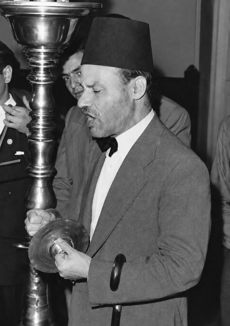 A man in a dark suit, bow tie and tarbush, holding a small cymbal, singing