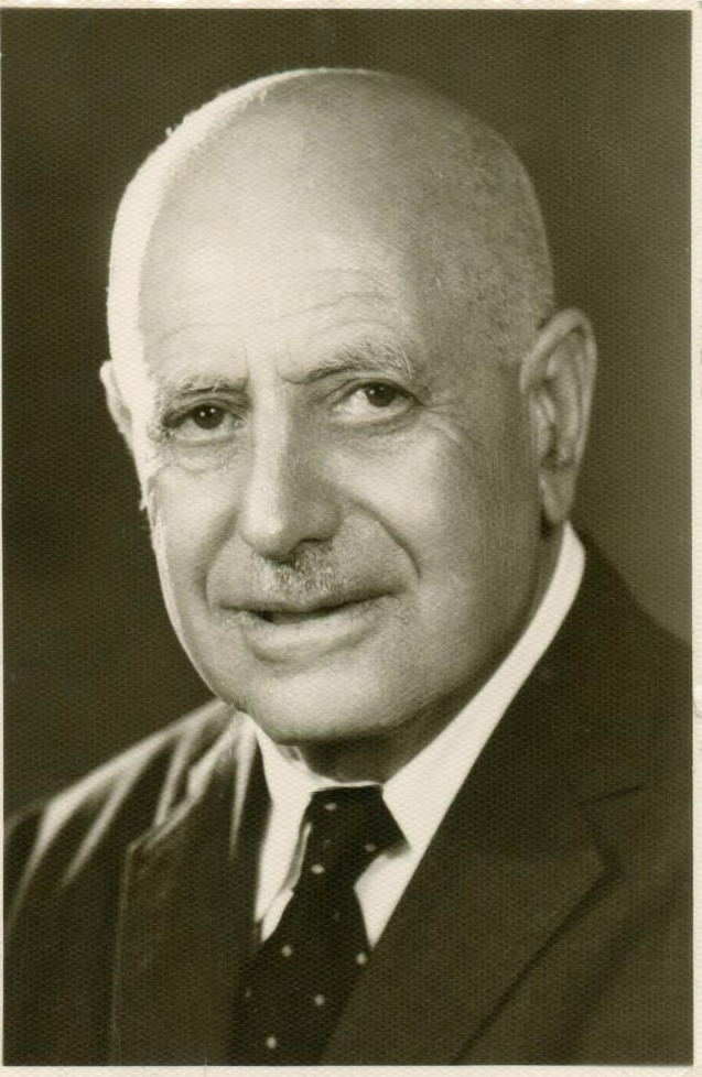 A bald, smiling man of about seventy in a dark suit and a dotted tie