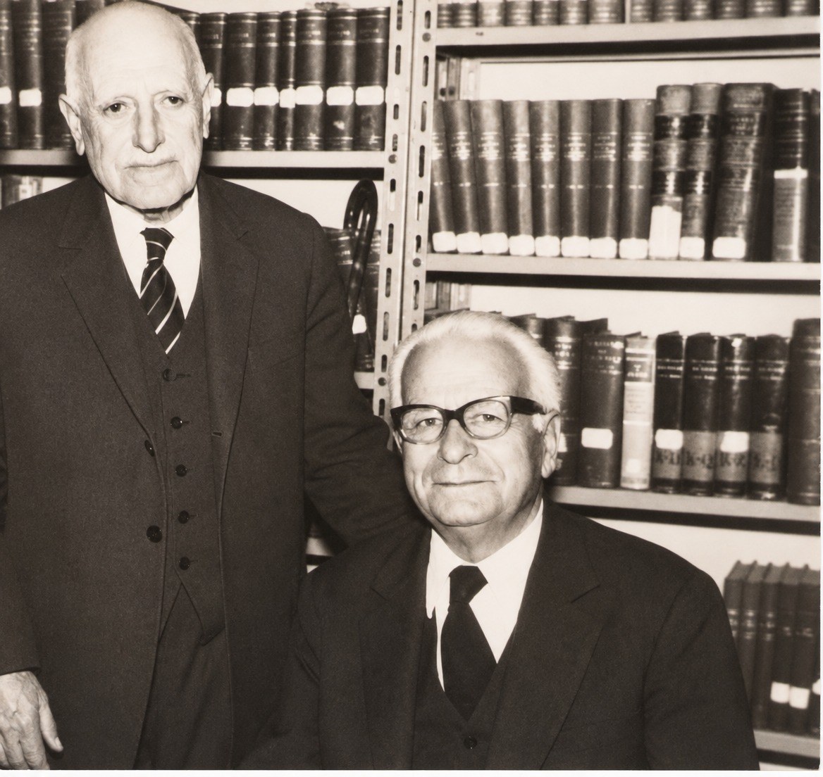 A tall old man standing in a three-piece suit beside a seated man in glasses, in front of shelves of bound volumes