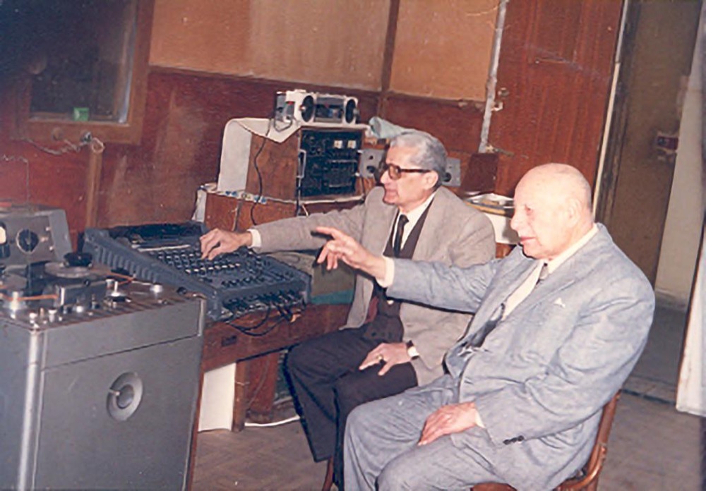 Two elderly men seated at a mixing desk beside a reel-to-reel tape machine
