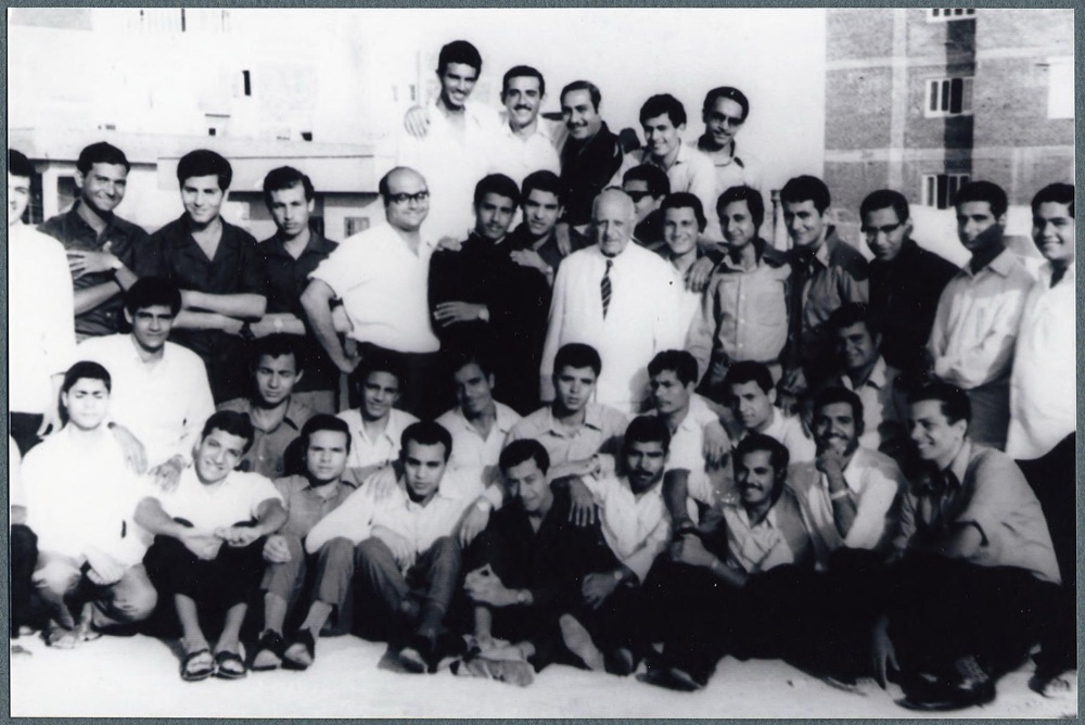 About forty young men, most in white shirts, gathered around an older man in a white suit