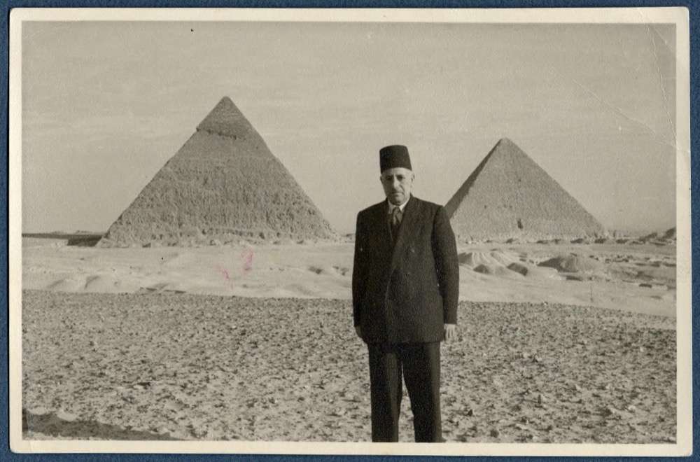 Ragheb Moftah, in a dark suit and a tarboosh, standing before two of the pyramids of Giza