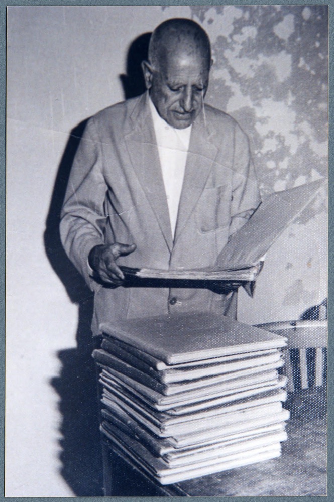 An elderly Ragheb Moftah, standing, turns the pages of a large bound volume from a tall stack of them