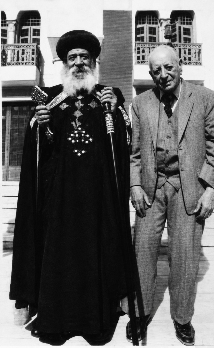 Pope Shenouda III in a black cope and turban, holding a hand cross and a staff, beside an elderly Ragheb Moftah in a grey three-piece suit