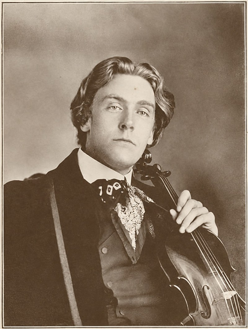 A young man with long wavy hair, in a dark coat and a patterned cravat, holding a violin