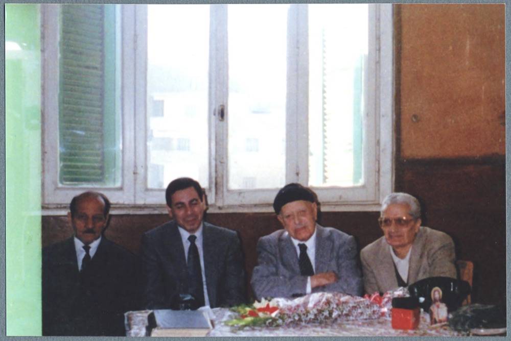 Four men seated at a table covered with flowers, in an office