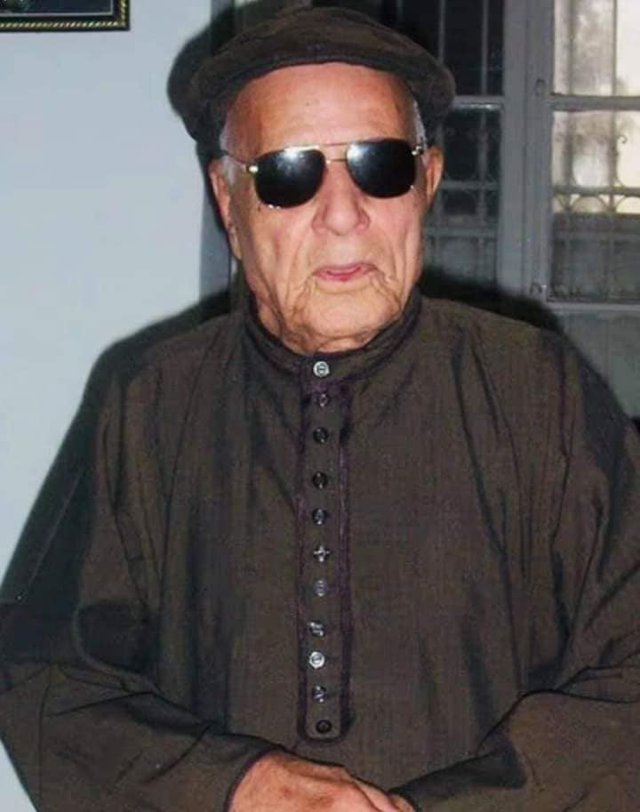 An elderly blind man in a cap, dark glasses and a brown robe buttoned to the neck