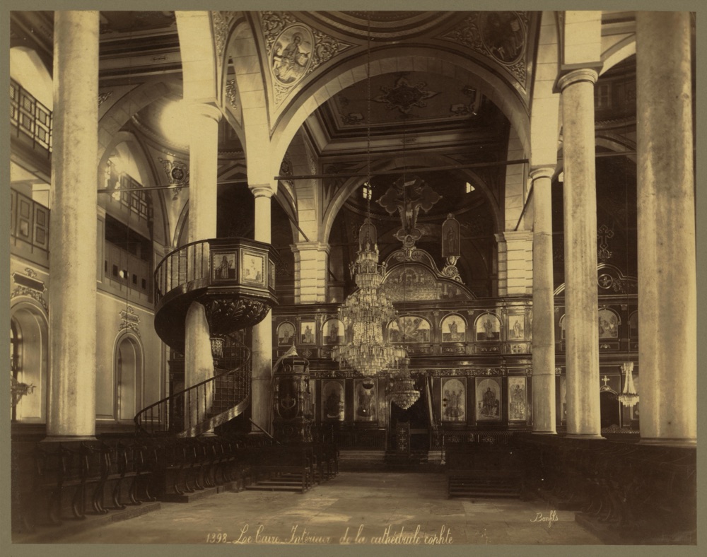 A large church interior with columns, hanging lamps, a spiral stair to a pulpit, and a carved screen at the far end